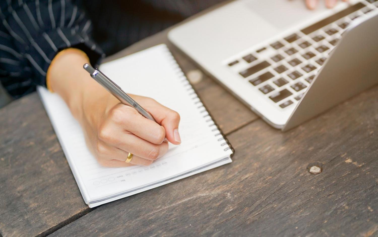 A person's hand writing in a notebook next to a laptop